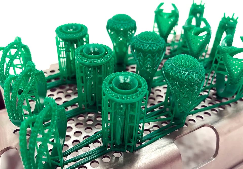 Green 3D-printed castable wax jewellery models on a perforated 3D printer build platform, ready for casting